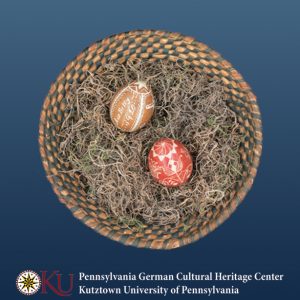 A green and orange woven basket is filled with grass and two carved eggs. The top egg is brown and the bottom egg is red.